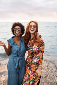 Two women laughing on the beach wearing dresses and sunglasses.